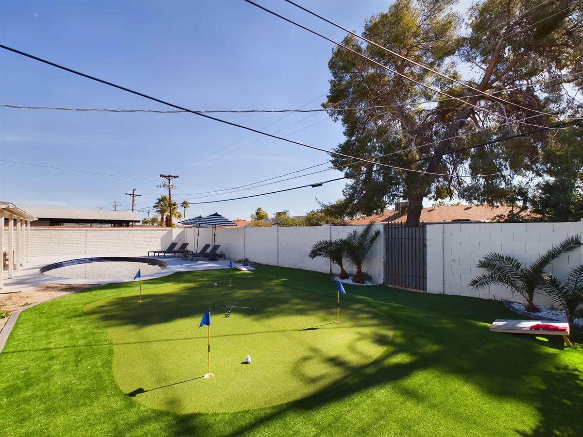 An exterior view of a backyard with a bright putting green, a glistening swimming pool, comfortable lounge chairs, and tall palm trees swaying overhead. The layout suggests a space designed for both relaxation and playful activities.