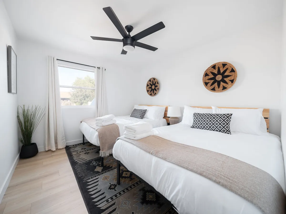 A bright and airy bedroom with two full beds dressed in white bedding, geometric patterned pillows, decorative wall hangings, a potted plant, and a window letting in natural light. The space is cozy, stylish, and ideal for families or groups.