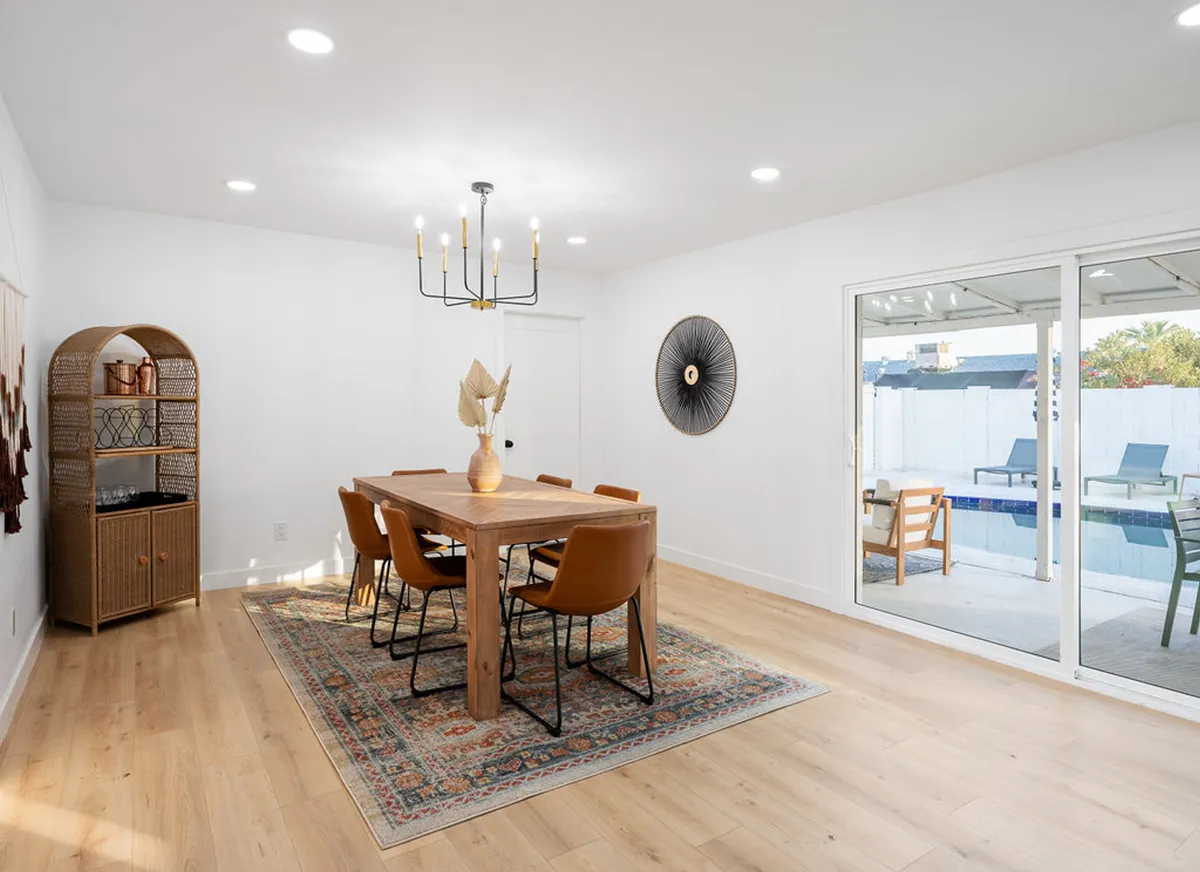 A warm, inviting dining area featuring a six-person table set beneath a chandelier. The area rug and rattan cabinet add layers of texture and style, while a sliding glass door offers views and easy access to the outdoors—suggesting a space where guests can linger over meals and conversation.