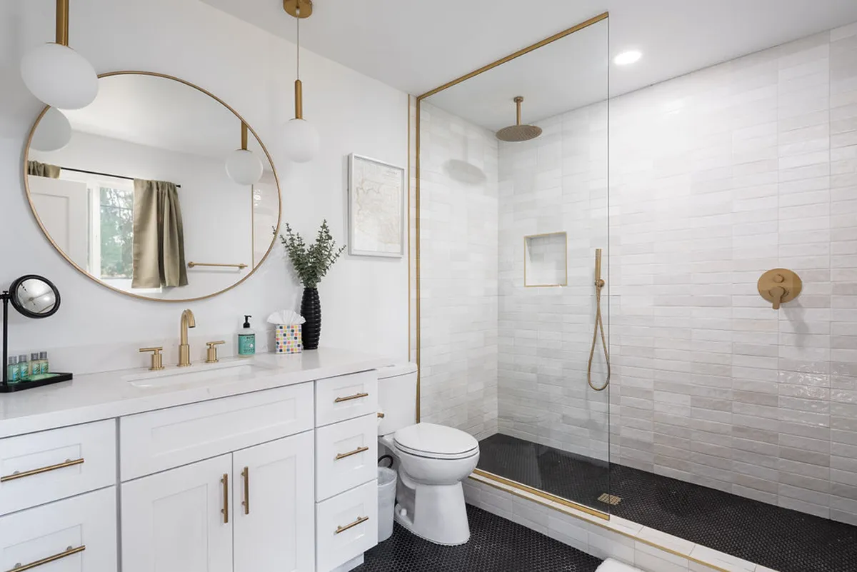 A spa-inspired bathroom featuring a large round mirror, glass shower enclosure, gold fixtures, and crisp white cabinetry. The mood is both serene and luxurious, suggesting a space designed for relaxation and rejuvenation.