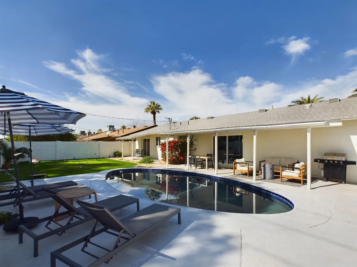 A bright, welcoming backyard scene featuring a sparkling swimming pool surrounded by lush palm trees and a neatly manicured lawn. The inviting patio area includes umbrellas for shade and ample space for socializing or relaxing. The setting is ideal for family fun and outdoor gatherings.