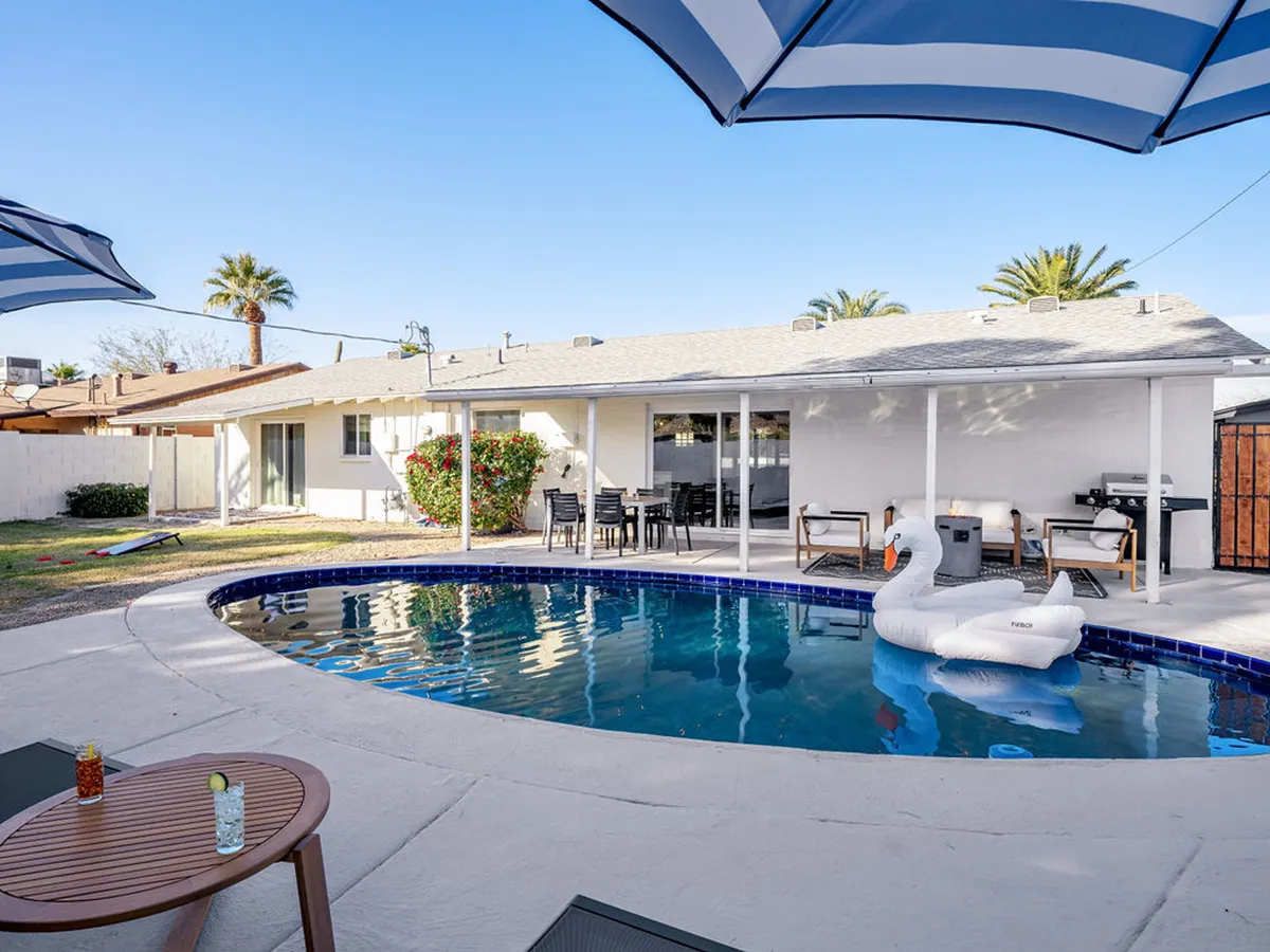 A vibrant backyard scene featuring a sparkling swimming pool with a floating swan, lush palm trees, and an inviting outdoor dining area in the background. Lounge chairs and subtle landscaping add to the resort-style ambiance.