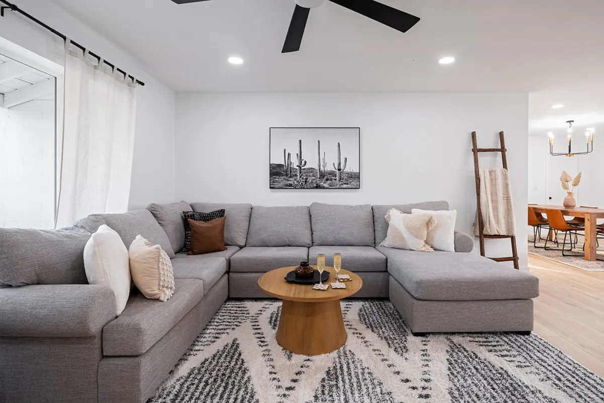 A cozy living room featuring a large gray sectional sofa, a wooden coffee table, textured throw pillows, and a framed cactus print. The space radiates comfort and is designed for relaxing, reading, or entertaining guests.