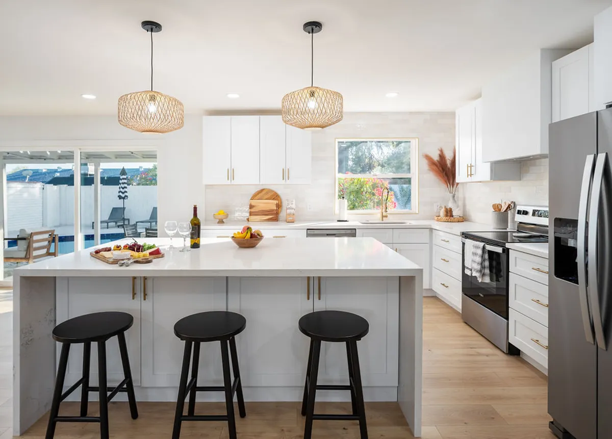 A warm, welcoming kitchen with a spacious island, stylish hanging light fixtures, a window offering an outdoor view, and decorative items like a fruit bowl, wine bottle, and glasses. The setup hints at both everyday convenience and special gatherings.