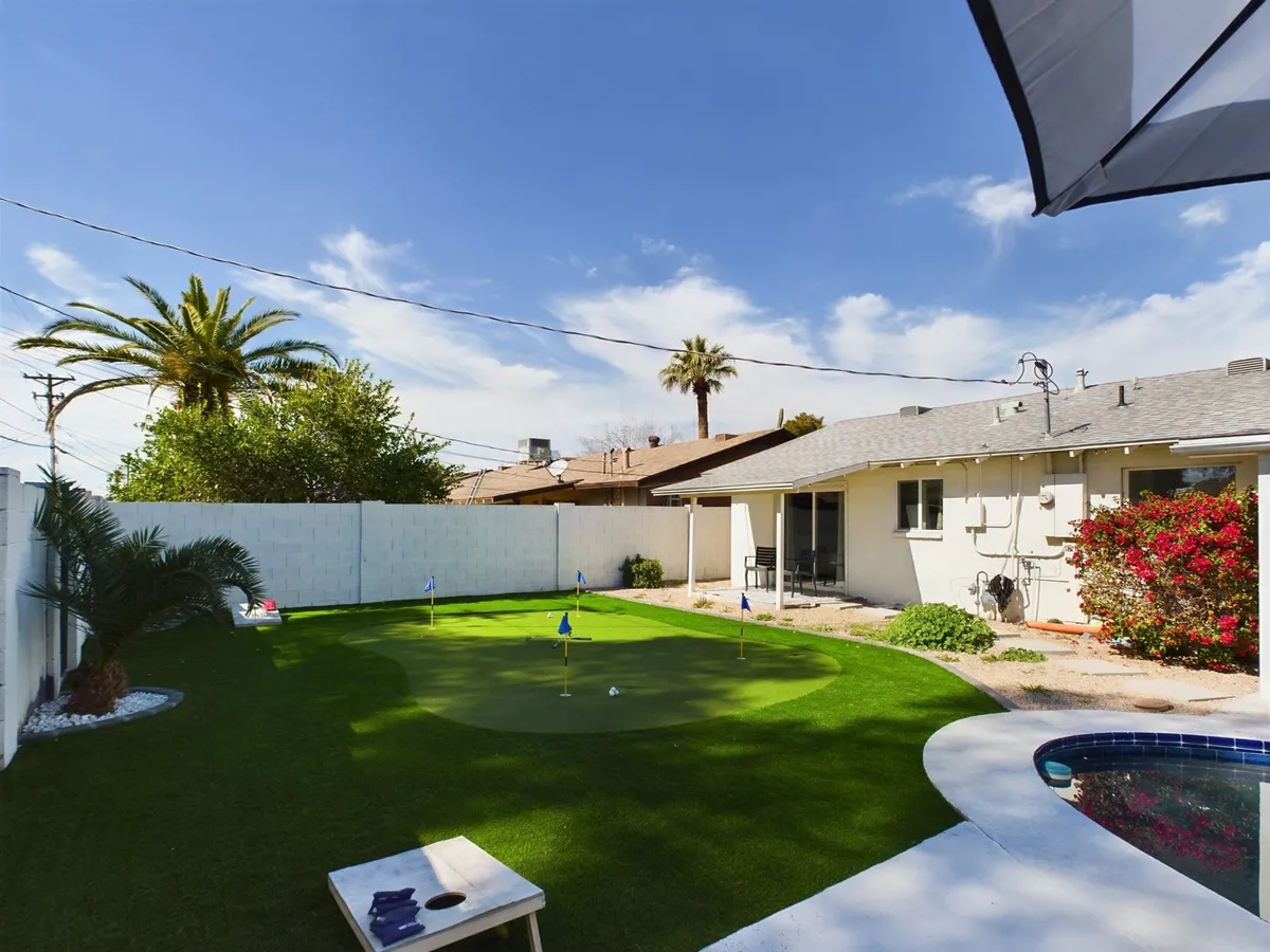 This exterior image showcases a lush backyard with a putting green, inviting jacuzzi, tall palm trees, and blooming shrubs. There’s a tranquil pool area visible, as well as a welcoming patio space ideal for gatherings and relaxing outdoors.