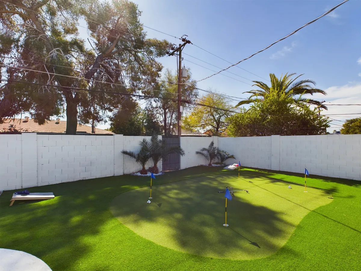 A playful backyard scene centered on a putting green framed by palm trees and a low white wall. The setting feels intimate, perfect for family gatherings or friendly competitions under the open sky.