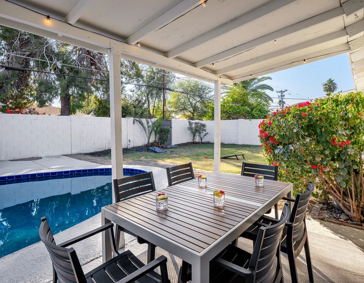 A cheerful exterior shot featuring an outdoor dining table next to a swimming pool and vibrant flowering bushes. The setup is ideal for alfresco meals and family gatherings in a beautiful, sunlit setting.