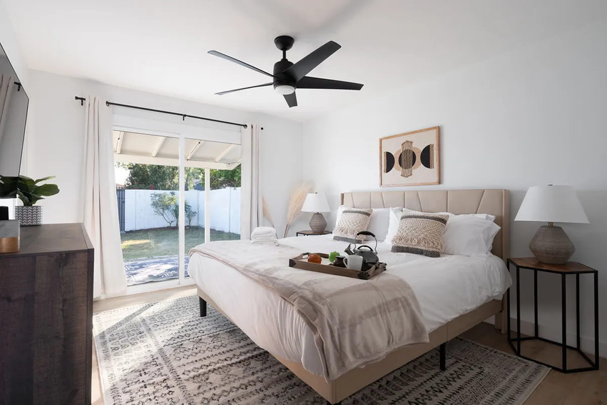 A serene bedroom featuring a plush bed with white linens, a breakfast tray, a large window allowing in natural light, a ceiling fan, and tasteful artwork on the wall. The space feels inviting and tranquil, ideal for restful mornings or quiet evenings.
