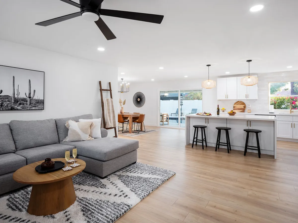 A bright open-concept space seamlessly connecting the living room, dining area, and kitchen. The contemporary gray sectional sofa creates a cozy gathering spot, while the modern dining table sits adjacent to the kitchen island with stools. Artwork featuring cacti adds a touch of local charm and vibrant personality.
