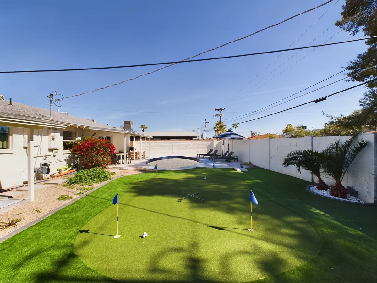 This image captures an inviting backyard with a putting green, sparkling pool, patio seating, and palm trees swaying in the background. The scene exudes relaxation and play, showcasing several recreational amenities for all ages.